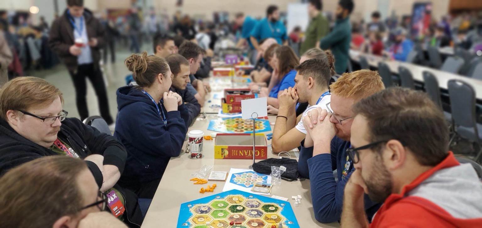 Players think carefully around a Catan board with many more games spread into the distance behind them.