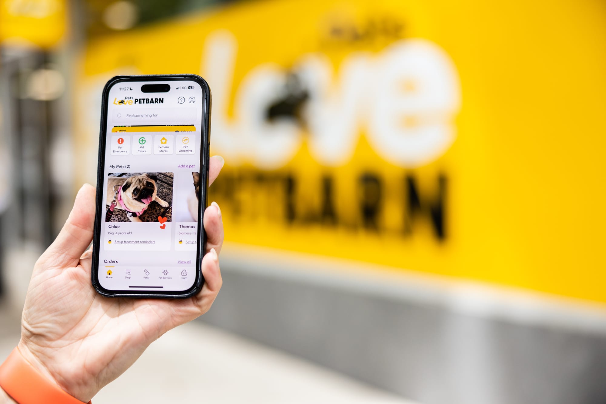 A hand holds an iPhone with the Petbarn app open with a Petbarn storefront blurred in the background.